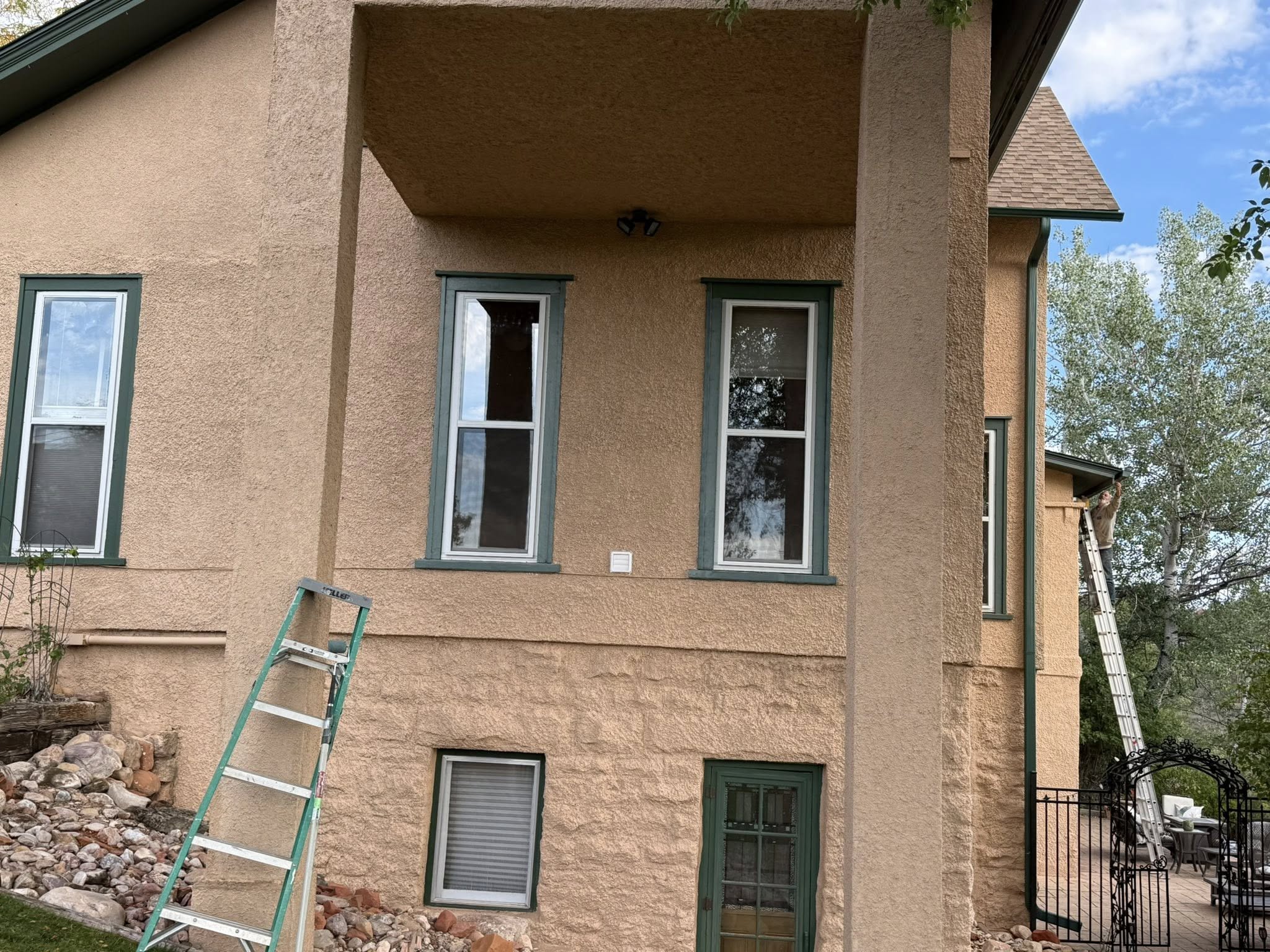Two-story tan stucco home with green window trim, exterior repaint in progress